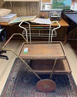 Photo of two vintage bar carts on wheels in a room with wood panel wall and desk nearby. One cart is metal painted white, missing two glass shelves; the other is wood and rattan with brass-colored metal frame and large wooden wheels.