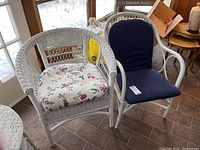 White wicker chair with removable floral cushion and painted bentwood chair with navy cushion side by side near a window.