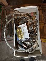 Box of assorted metal hardware parts including lamp fittings, drawer handles, decorative rings, placed on a surface.