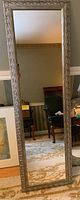 Full view of the full-length mirror showing the entire rectangular shape and reflections in the glass. The mirror stands vertically against a wall and some furniture is visible reflected.
