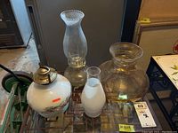 Lot of glass oil lamps and glass shades on display on a shelf with surrounding items.