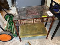 Metal Asian-style side table showing geometric patterned metal frame with glass top and bottom shelf. Worn metal finish with rust and discoloration visible.