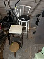 Three stools shown together. Tall white metal bar stool with black vinyl seat, vintage metal-legged kitchen stool with off-white vinyl cushion, and small round wooden stool.