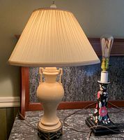 Photo showing two table lamps side by side on a surface, the taller lamp with cream ceramic and metal lattice base with pleated beige shade, the shorter lamp with floral ceramic and wood base without a shade.