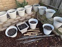 Overview of all 13 planters arranged on stone ledges and ground, some containing soil, others empty. Bamboo sticks and wooden pieces placed in front.