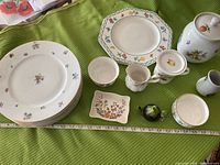 Photo of floral chinaware set including plates, bowls, pitchers, cups, and jars on green fabric with measuring tape for scale