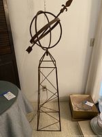 Full view of the tall rust-colored wrought iron weather vane sculpture showing the base, tower, globe, and arrow.