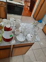 Top-down photo of the countertop showing stacked white mugs, etched glasses, espresso cups on saucers, and the vintage thermos along with additional saucers.