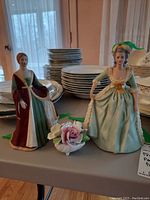 Two Franklin porcelain figurines and one porcelain floral piece displayed on a table with background of stacked dishes. The figurines show historical female figures in elaborate dresses.
