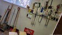 Wood-handled garden tools including various shovels, rakes, brooms, snow shovel, hanging on wall and leaning near window
