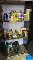 Cabinet interior with three shelves showing assorted cans, bottles, tubes, and boxes including Maxwell House can, Martin's apple juice can, and hardware supplies.