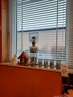 Photo showing the decanter, five shot glasses, wooden house and angel figurine on a windowsill with blinds.
