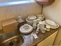 Photo showing the full set of dishes, cups, saucers, and service pieces arranged on a countertop near sink.