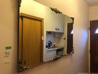 Front view of large rectangular hallway mirror with decorative ornate corner trim, showing reflection of wooden door and hallway.