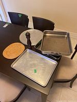 Photo showing two rectangular steel oven pans, a round pizza tray, a baking sheet, a wooden pepper mill, and woven trivet arranged on a dining table.