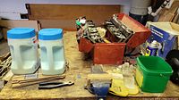 Photo showing the open red metal tool box filled with hardware and various hand tools, along with bowls, plastic containers of ice melter, stain bottles, and a blue steel bench vice on a wooden workbench.