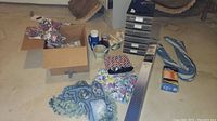Photo showing assorted items on the floor including box with floral curtains, thermoses, mugs, blanket, and plastic storage drawers