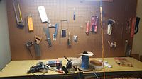Wide view of assorted hand tools hung on pegboard including saws and hand tools along with workbench holding soldering kit, wire spool, and hand plane.
