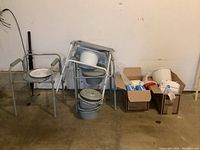Three gray portable potty chairs with removable buckets, one shown with lid, two stacked. Two boxes containing adult disposable underwear and hygiene supplies on the floor nearby.