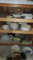 Wooden shelf containing various Pfaltzgraff bowls, white Corningware bowls, baking dishes, and other kitchenware including plastic containers and a folding colander.