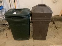 Front view of two outdoor plastic trash cans, one dark green Rubbermaid and one brown with handle and wheels, resting on a concrete floor.