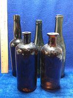 Front view of 5 antique glass bottles standing upright on blue surface with a ruler for size reference. Includes dark brown and green glass bottles, some with visible embossing and one with chipped lip.