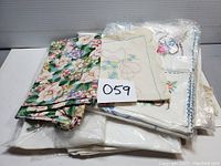Photo showing stacked assorted linens including one with bright floral prints and several embroidered towels.