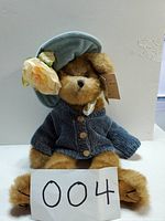 Full front view of the plush bear sitting upright with tag attached, showing clothing and detailed flower on hat.
