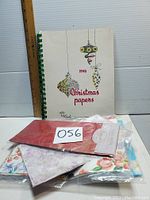 Vintage 1958 Christmas gift wrap sample book next to a ruler and several sheets of colorful vintage wrapping paper in plastic packaging on a white surface.