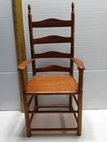 Front view of wooden doll chair with ladder back, armrests, and seat, standing against white panel background with wooden ruler for scale.