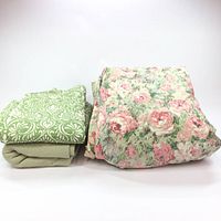 Photo showing one thick floral comforter folded on the right, and a stack on the left with a green blanket with white pattern on top and a light green comforter underneath.