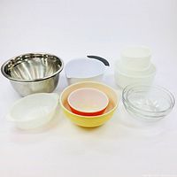 Lot arranged showing all 9 bowls from various materials: stainless steel, clear glass Pyrex, white opaque plastic, yellow and orange plastic. The diversity of sizes and slight damage visible such as chips can be seen.