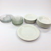 Photo showing multiple stacks of white ceramic plates and bowls alongside stacks of clear glass plates, highlighting embossed grapevine pattern and quantity.