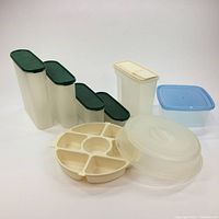 Photo showing four tall vertical Tupperware containers with green lids alongside one white rectangular container with beige lid, one small blue-lid container, and a cream round multi-compartment tray with lid.
