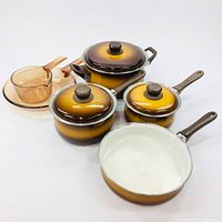 Photo of entire set showing four vintage enamel cookware pieces with lids and two amber glass dishes with lids on white background.