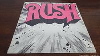 Front cover of Rush self-titled vinyl LP album with large pink RUSH logo and white background splattered with black and gray lines.
