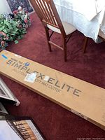 Long unopened cardboard shipping box labeled 'Simplex Elite' on red carpet alongside furniture and floral arrangement
