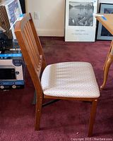 Side view of wooden folding chair with light fabric seat and vertical slat back on burgundy carpet.