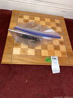 Photo showing the wooden chess board with light and medium brown squares, flat on a carpet with the chess pieces in zip bags placed on top.