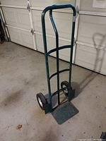 Green metal hand truck dolly shown upright with visible frame, wheels, and toe plate