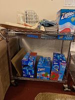 Metal rolling cart with household items on shelves including Ziploc bags and cloths.