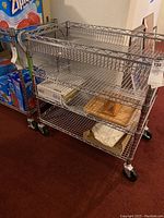Front angle view of chrome metal 3-tier rolling cart with items on shelves and Ziploc box in background.