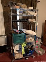 Full view of metal shelving unit loaded with various textiles, pillow, and plastic storage bins.