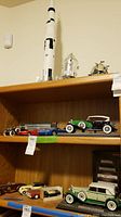 Three-shelf wooden shelving with assembled spacecraft models on top shelf, diecast cars and train set in middle, additional diecast cars on bottom
