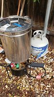 Full view of Brinkman propane deep fryer with lid, burner stand, gas hose, and Blue Rhino propane tank beside it on ground covered with leaves.