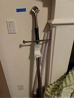 Silver-tone decorative wall sword leaning against wall, showing crossguard and faux leather grip