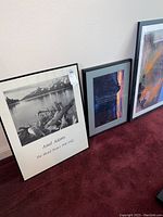 Three framed art pieces leaning against wall on carpet: Ansel Adams black and white print, sunset mountain color photo, and large abstract piece