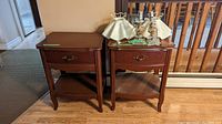 Two reddish-brown wooden side tables each with a single drawer, lower shelf, and curved legs. Two lamps with fabric shades on top of tables.