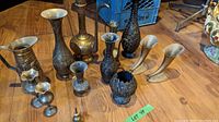 Photo showing 10 black metal vases of varied sizes and shapes with floral and ornate decorative patterns, placed on wooden floor.