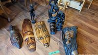 Photo showing the full lot of seven wooden carved items including five masks and two figurine sculptures on a hardwood floor.